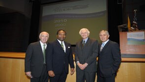 Four men, all in ties and dark suits, stand close and face the camera, smiling.