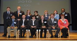 Thirteen people appear, with three women and three men sitting on chairs in the front row and four women and three men standing behind them in the back row, all smiling for a posed picture.