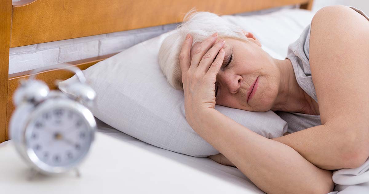 Older woman in bed trying to sleep with a metal alarm clock in the foreground