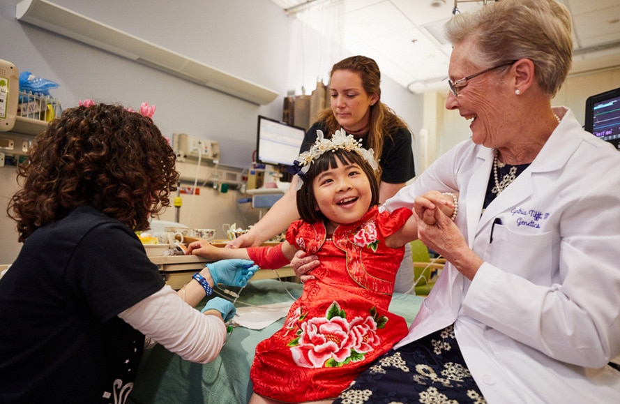  A child with GM1 gangliosidosis in a red dress sitting on a bed with three health care professionals holding up her arms as she takes part in a clinical trial at the NIH Clinical Center.