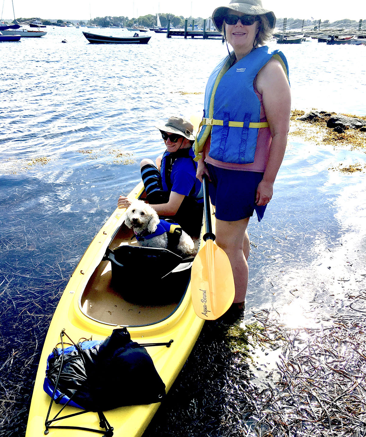 Teresa Jones standing in shallow water next to a yellow kayak with a friend and small dog inside it