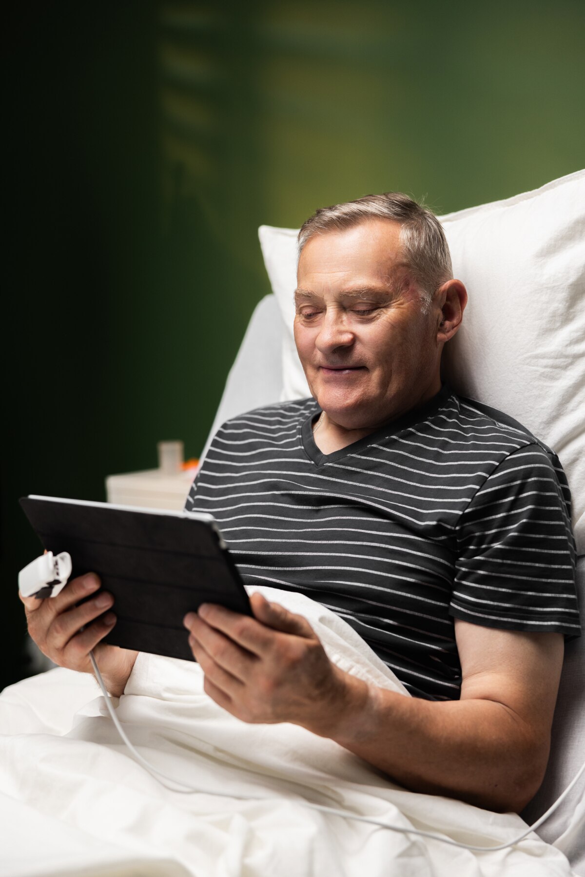 An older male sitting in a hospital bed takes part in online behavioral therapy through an electronic tablet.