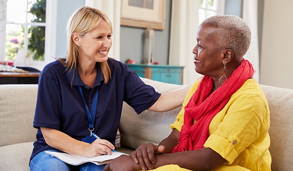 Image of a young female doctor sitting on a couch smiling, while holding a clipboard, with one arm on the shoulder of a smiling middle-aged patient. 