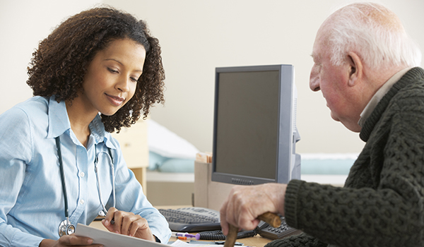 Image of female doctor sitting at a desk, holding papers and speaking to an older man with a cane.