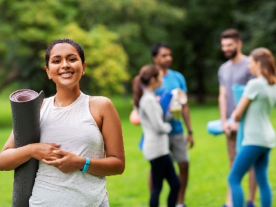 A woman smiling holding a yoga mat with a group of friends in the background. 