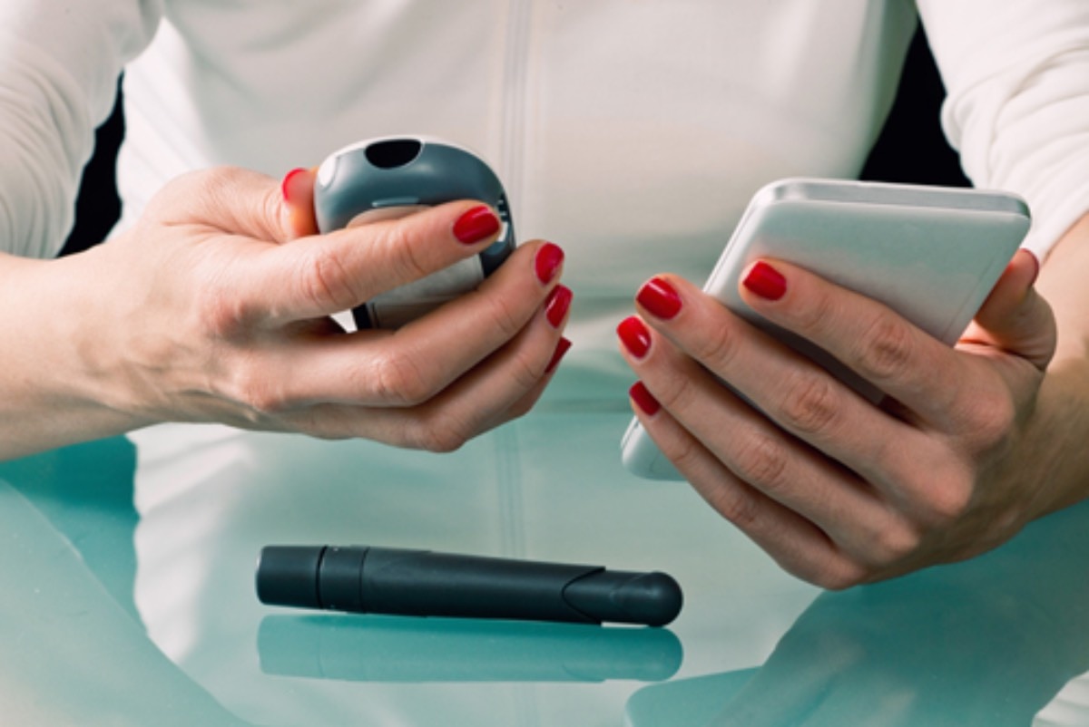 A woman compares her blood glucose reading from a standard glucose monitor with a CGM reading on a smartphone. A lancet is on the table in front of her.