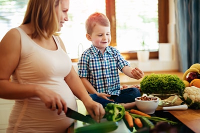A pregnant woman chopping vegetables in the kitchen with her young son.