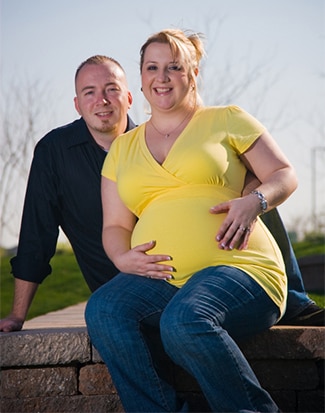 A pregnant woman sitting outdoors with her husband behind her.