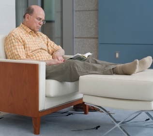 Photo of a man sitting reading a book with his feet resting on footstool.