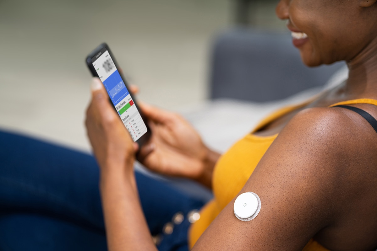 A woman with a smart sensor attached to her arm looks at a mobile phone that shows her blood glucose reading.