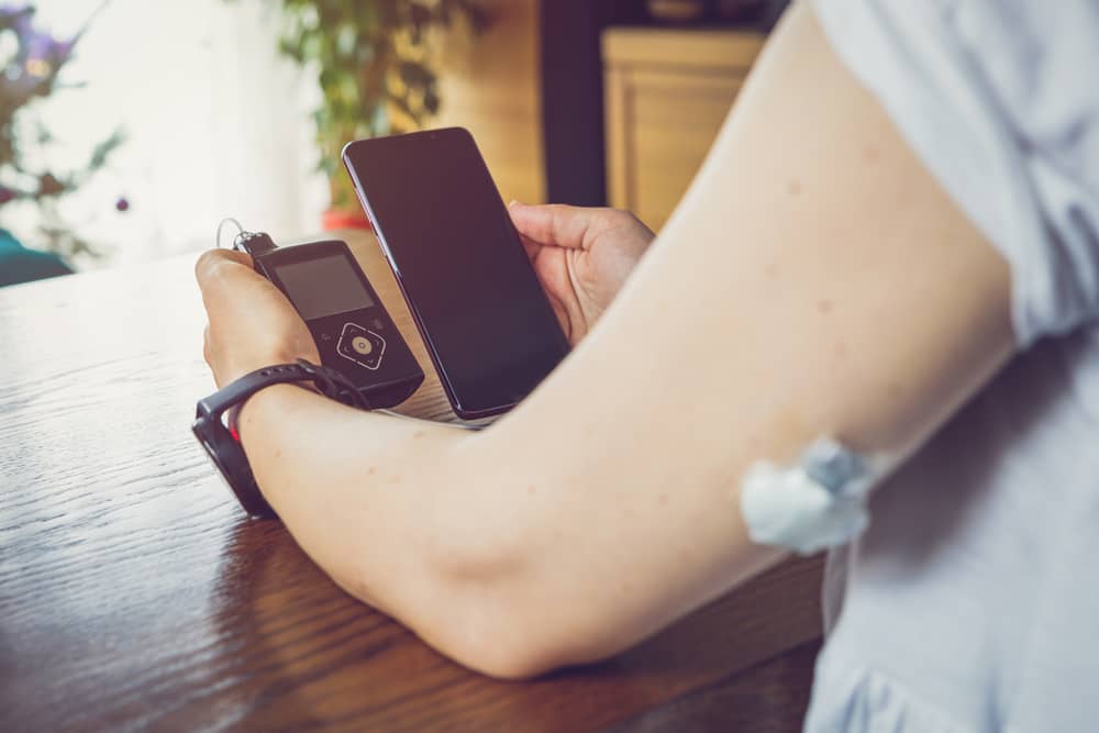 Una mujer está sentada en una mesa con un teléfono inteligente y un monitor continuo de glucosa, que está conectado a una aguja insertada en la parte posterior de la parte superior del brazo.