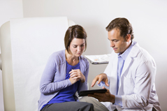 Male healthcare provider in white lab coat holding a clipboard talking with a female patient.