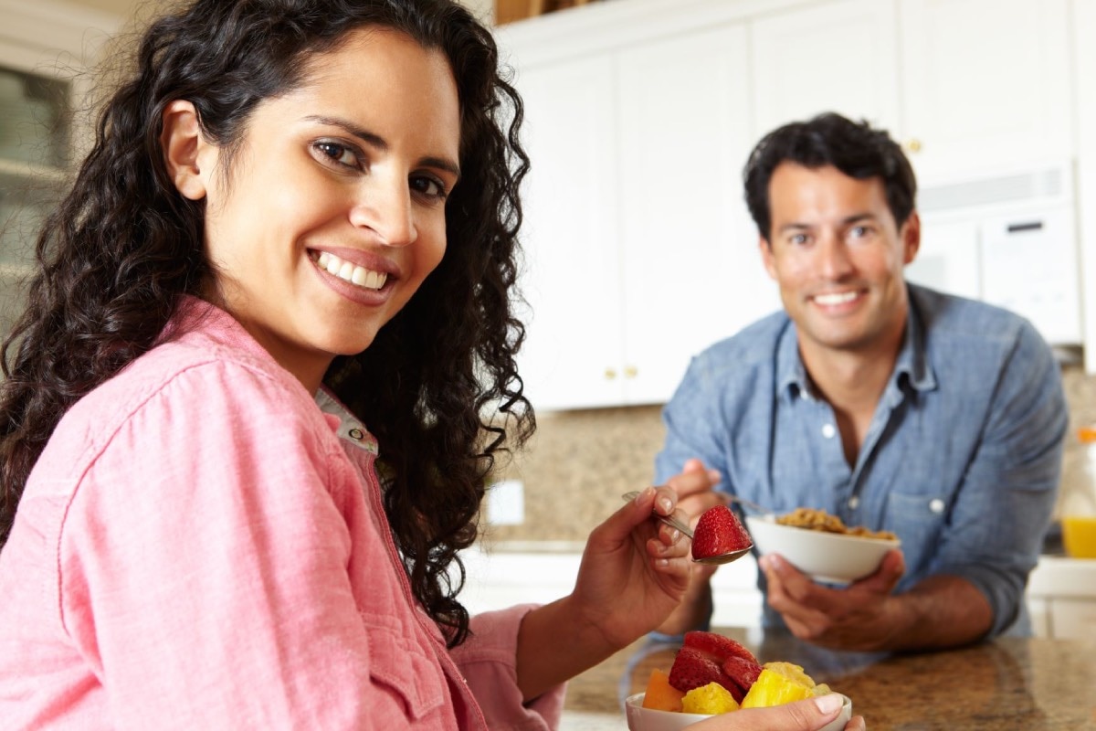 Mujer comiendo un plato pequeño de fruta y hombre comiendo un plato pequeño de cereal.