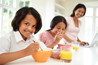 Two children eating breakfast with their mother in the background.