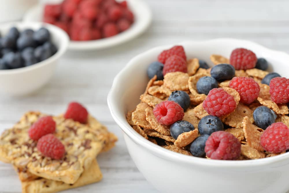 Un tazón con cereal integral y bayas frescas al lado de dos galletas ricas en fibra y frambuesas.