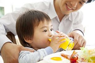 A young child drinks a glass of orange juice with help from his father. A plate of eggs, bacon, and vegetables sits on the table in front of them.