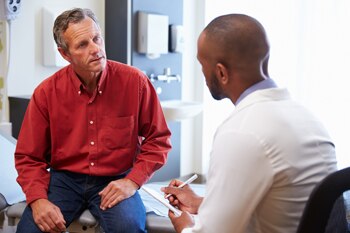 Photo of a man sitting on an examining table talking to a male doctor sitting in a chair.