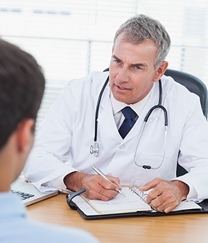 A male doctor sitting at desk writing a prescription for a male patient.