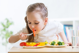 Niña comiendo un tomate con pimientos amarillos, brócoli, zanahorias y pasta. La foto también muestra un vaso de agua.