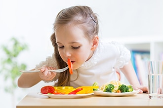Girl eating a tomato with yellow peppers, broccoli, carrots, and pasta. Photo also shows a glass of water.