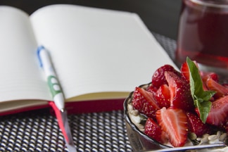 Un bolígrafo y un diario abierto junto a un bol con fresas y cereal.