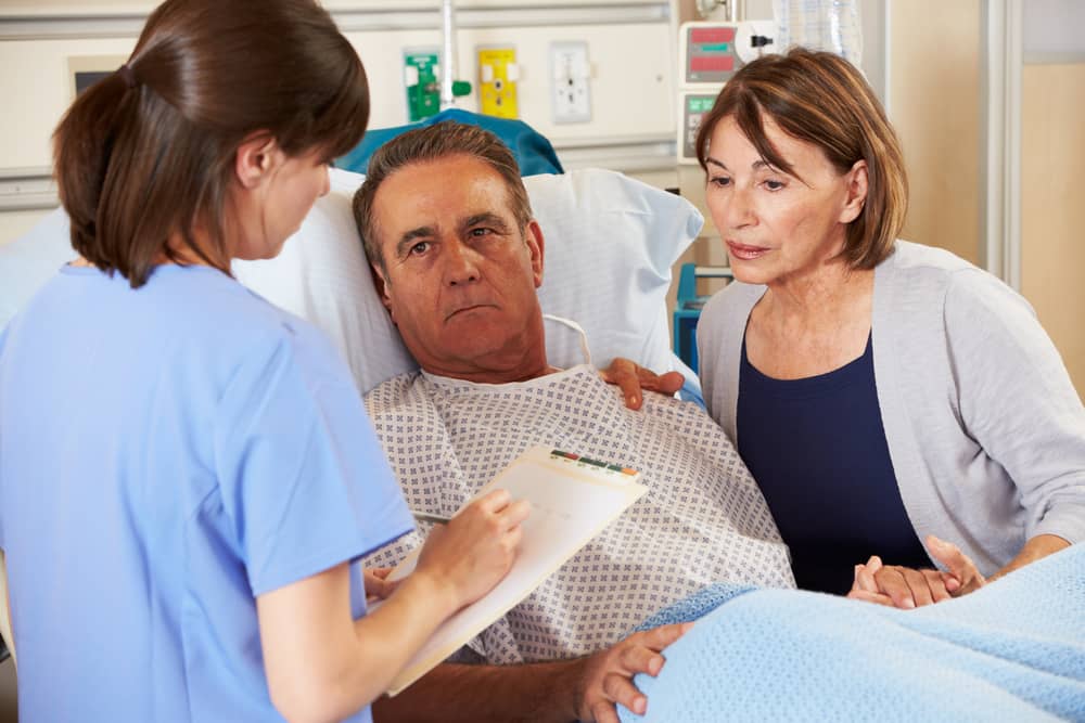 Health care professional taking notes and talking with a patient in a hospital bed and his spouse.