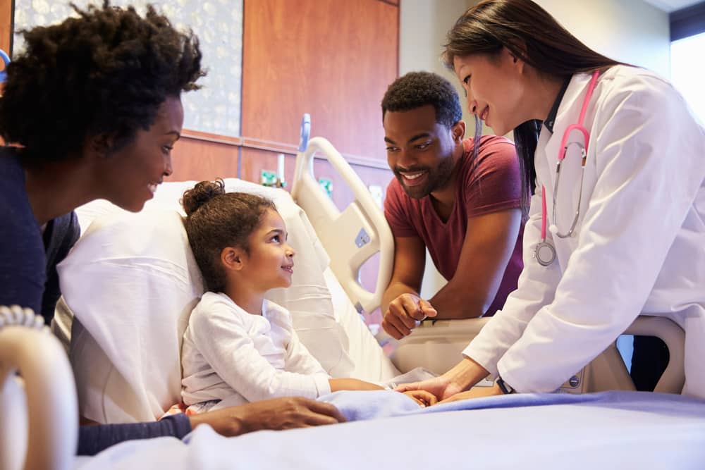 A child sits up in a hospital bed, surrounded by her parents and doctor.