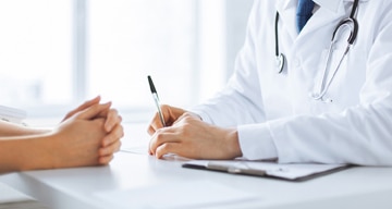 Close up of a doctor’s desk. On one side, a doctor is writing a note. A patient’s folded hands rest on the other side of the desk.