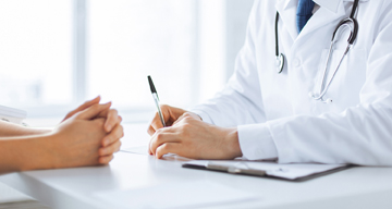 Doctor writing data on a clipboard while sitting in front of a patient with folded hands.