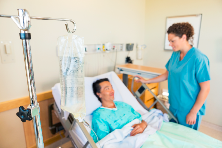 A man in a hospital bed with an IV solution bag hanging from a metal pole and a health care professional standing by his bed.