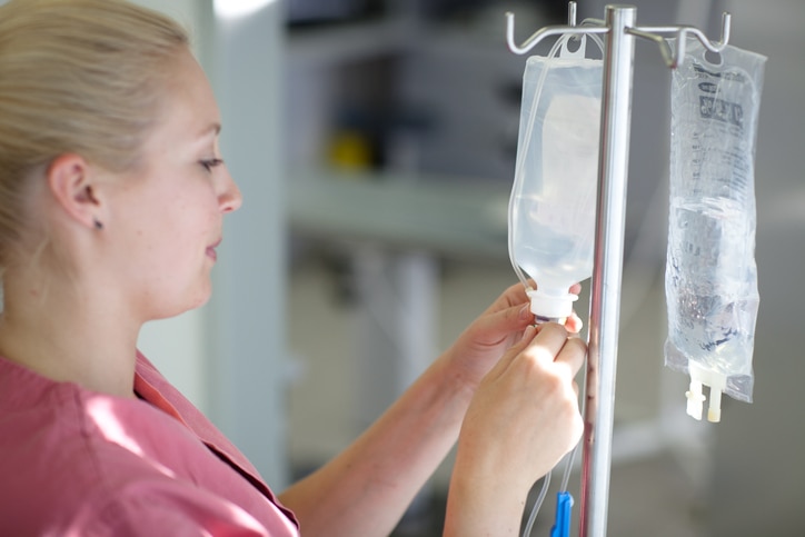 A health care professional holds the opening of an IV drip bag, which hangs from an IV pole.