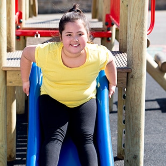 A young girl with overweight or obesity who is sliding down a slide.