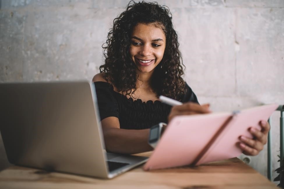 Female college student looks at a laptop computer screen while writing in a notebook.