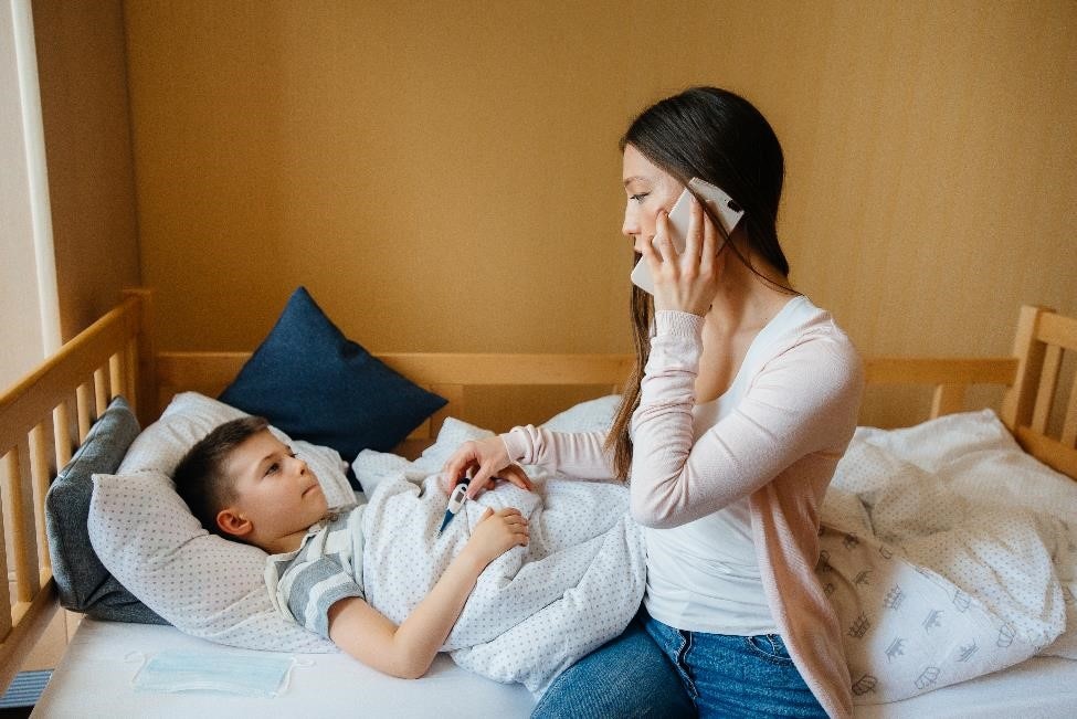 Madre con el termómetro en la mano sentada al lado de una niña enferma y llamando al médico por teléfono