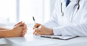 Doctor que escribe los datos en un portapapeles mientras está sentado frente a un paciente con las manos juntas.