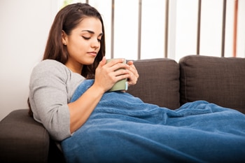 Una mujer joven que está recostada en un sofá envuelta en una cobija y sosteniendo una taza grande.