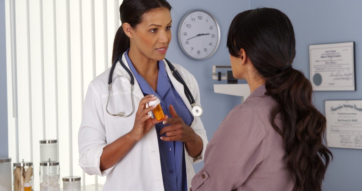 Una doctora habla con una paciente y sostiene un frasco de un medicamento recetado.