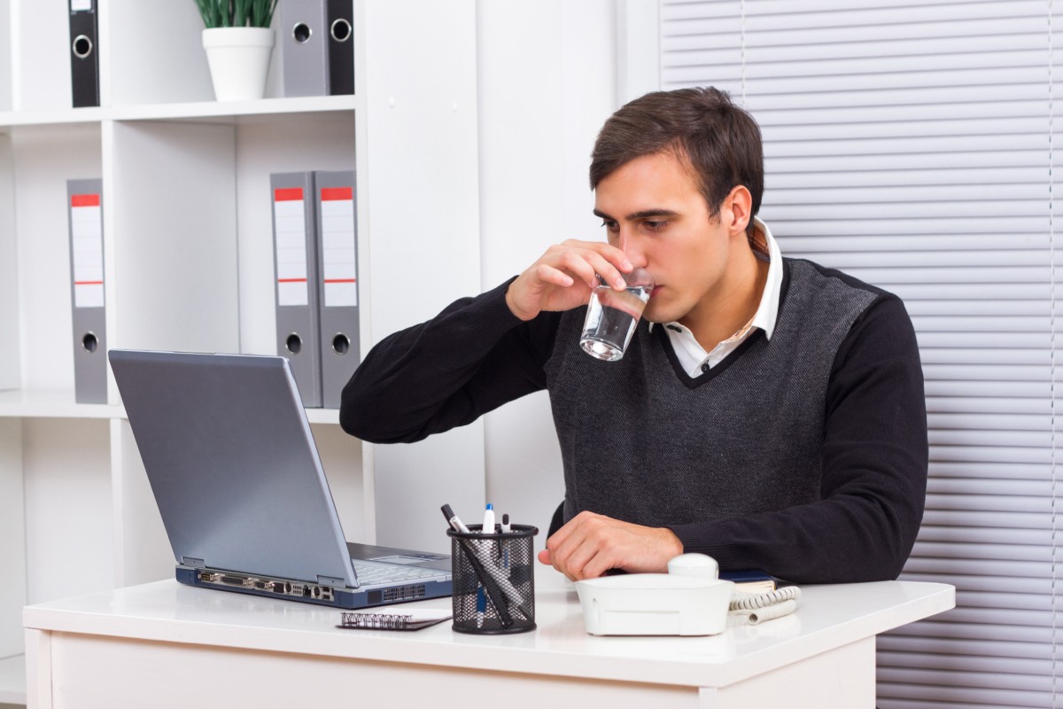 Un hombre sentado en un escritorio bebe un baso de agua.