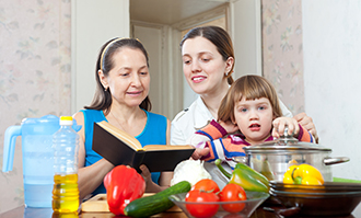 Fresh vegetables, cooking oil, water, and a large pot are in front of two women looking at a book. A young child reaches for the lid of the pot.
