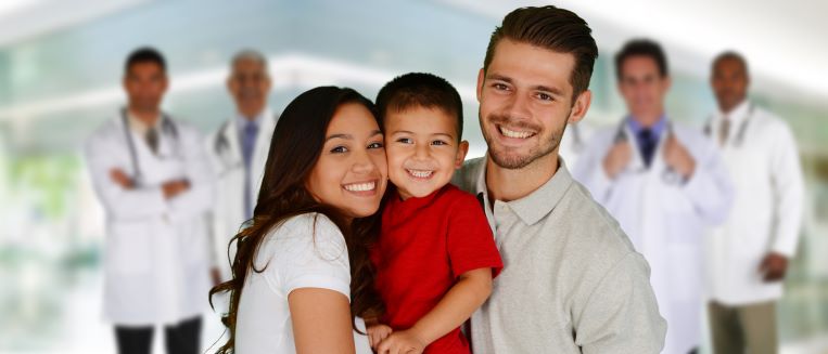 Parents standing and holding a young boy between them, with a team of health care professionals looking on in the background.