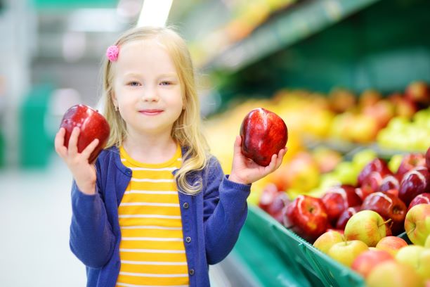 A young girl standing in the fruit section of a grocery store holding a red apple in each hand.