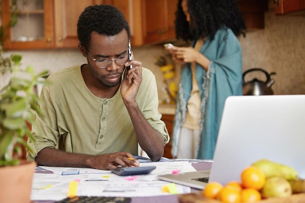 Man sitting at a kitchen table with a calculator, reviewing bills, and talking on the phone.