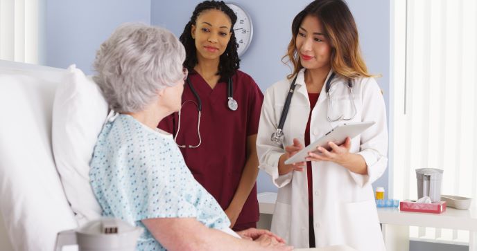 Two health care professionals talking with an older patient who is sitting in a hospital bed.