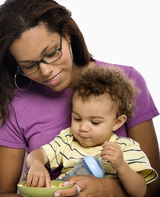 Mother holding a toddler who is holding a bottle and eating food from a bowl