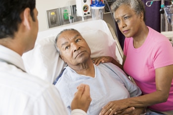 Ein älterer Mann in einem Krankenhausbett mit seiner Frau und seinem Arzt an seiner Seite.