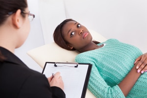 Photo of a woman who is reclining in a doctor’s office and looking at a health care professional who is taking notes.