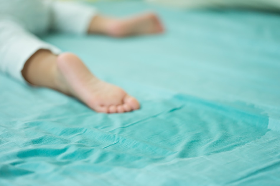 A child in pajamas lies near a wet spot on the bed.