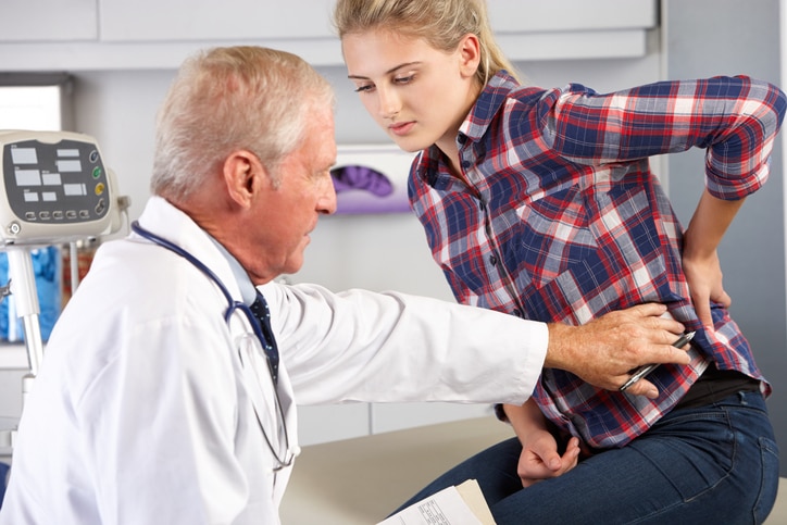 Teen girl talks to doctor and holds a hand to her lower back.