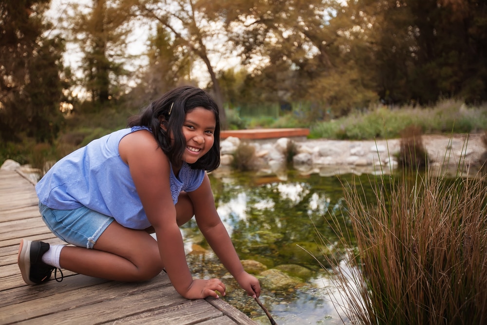 Girl playing outdoors, kneeling on a wooden pathway over a pond.