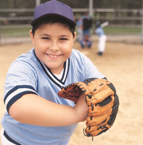 A smiling boy in a baseball uniform holds a ball and baseball glove.